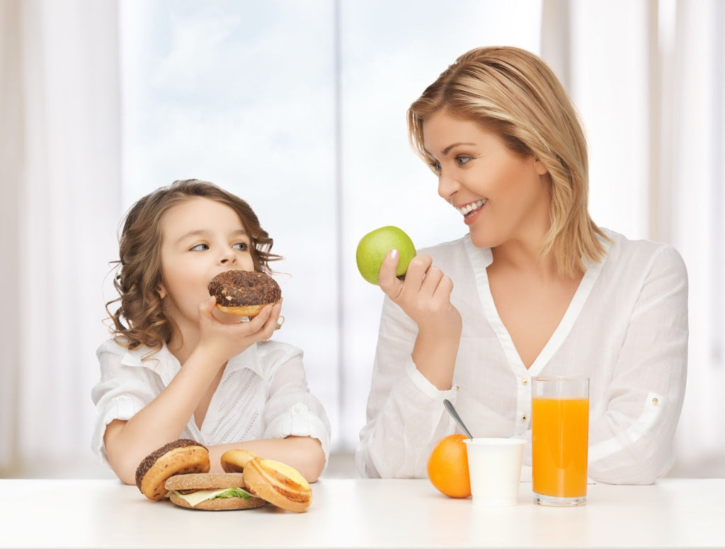 Mother and child sitting at a table. Kid has unhealthy food in front of him while mother holds an apple. 