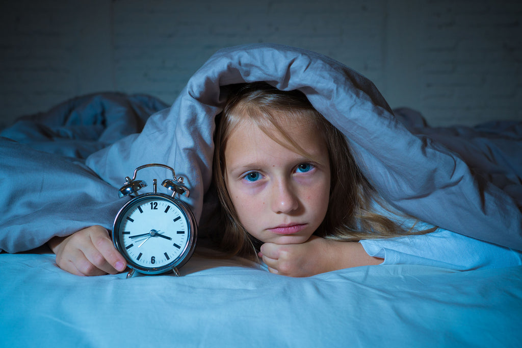 Cute little girl with Autism who can’t fall asleep and is holding an alarm clock with the blankets over her head.